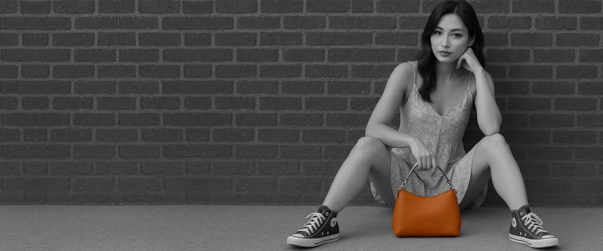 Woman sitting on the ground holding an orange handbag against a brick wall.