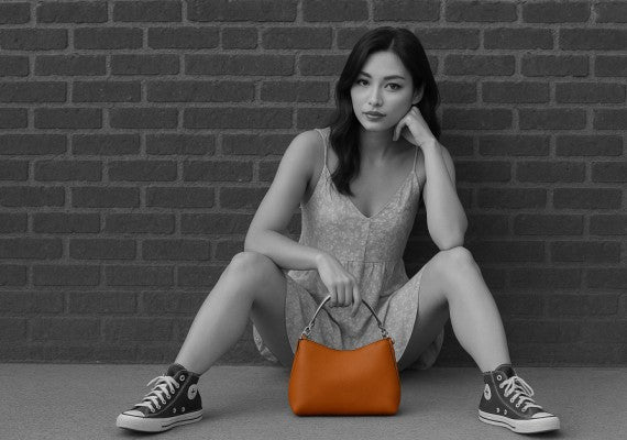 Woman sitting on the ground holding an orange handbag against a brick wall.