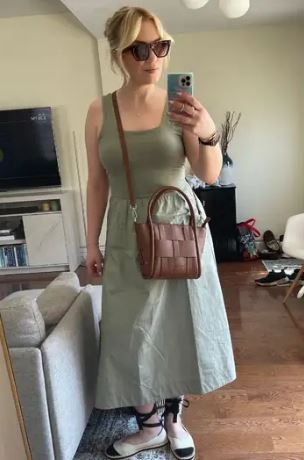 Woman taking a mirror selfie wearing a green dress and brown vegan handbag in a living room.